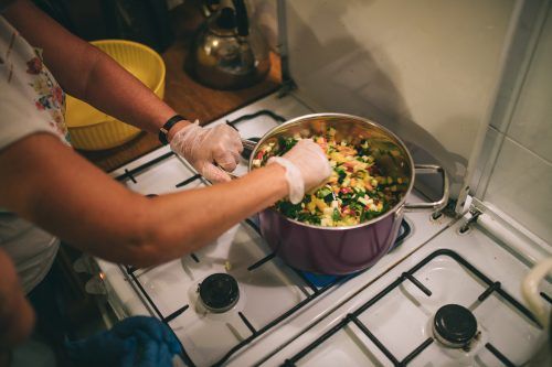 Widok z góry na gazową kuchenkę, na której stoi garnek wypełniony pokrojonymi warzywami i ziołami, widać ręce osoby, która miesza produkty w garnku