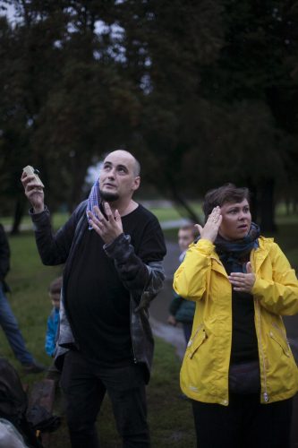 Łysy mężyczyzna pokazuje ręką niewidoczny na zdjęciu punkt na niebie, stojąca obok niego kobieta tłumaczy jego słowa na Polski Język Migowy. Mężczyzna ubrany jest w czarną bluzę, kobieta w żółtą kurtkę przeciwdeszczową.
