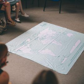 Mapa świata rozłożona jest na podłodze, grupa ludzi obserwuje ją siedząc na krzesłach
