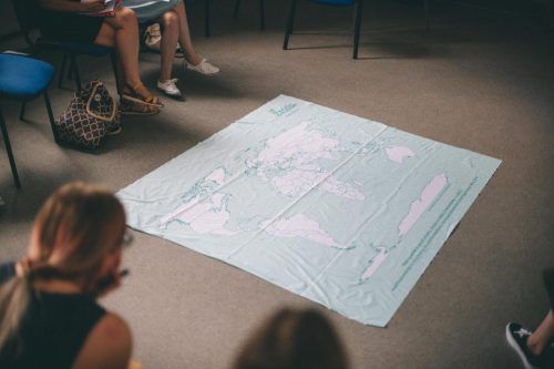 Mapa świata rozłożona jest na podłodze, grupa ludzi obserwuje ją siedząc na krzesłach