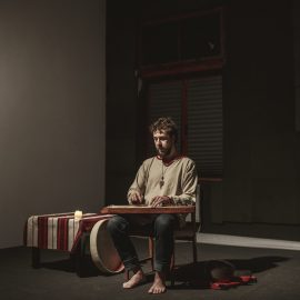 Mężczyzna siedzi w ciemnym pomieszczenu na krześle. Nie ma butów, ubrany jest w szarą koszulę i jeansy. Na kolanach trzyma instrument, na którym gra. Obok niego jest stolik z białoczerwonym obrusem i świecą.