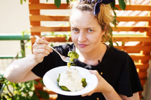 Zdjęcie, Aleksandra w trakcie jedzenia brokułów z białym sosem. Jest na zewnątrz i trzymając widelec patrzy się prosto w obiektyw.