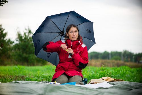 Zdjęcie kobiety siedzącej na kocu na trawie. Kobieta ma na sobie czerwoną kurtkę i w ręce trzyma parasolkę.
