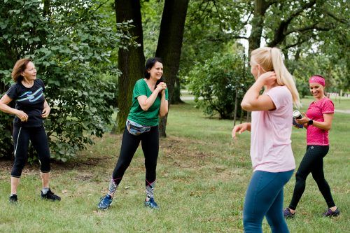 Cztery kobiety ćwiczą w parku. Rozciągają mięśnie rąk. Wszystkie są uśmiechnięte - widać, że treningi sprawiają im dużo radości
