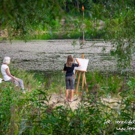 Zdjęcie, dziewczynka stoi na polanie i maluje na płótnie jezioro znajdujące się przed nią. Obok niej na kocu siedzi starsza kobieta.
