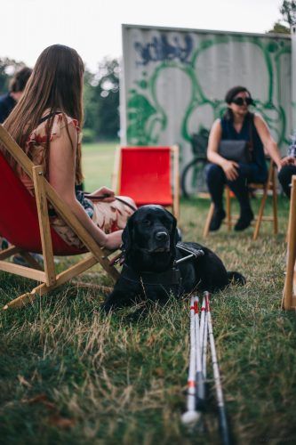 Pies przewodnik leży na trawie obok swojej właścicielki, która siedzi na leżaku. Przed nim znajduje się biała laska. Pies to czarny labrador.