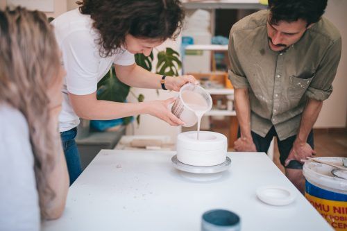 W pracowni ceramicznej dwie osoby nachylaja się nad stołem wlewając masę do formy odlewniczej, z której powstanie ceramiczny kubek.