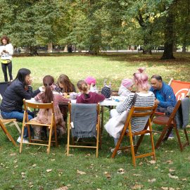 Zdjęcie wykonane w plenerzee w Parku Południowym. Na zielonej polanie przy rozstawionych stołach siedzą uczestnicy i uczestniczki warsztatów tworzenia bajek.