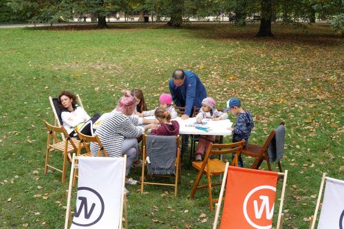 Zdjęcie wykonane w plenerzee w Parku Południowym. Na zielonej polanie przy rozstawionych stołach siedzą uczestnicy i uczestniczki warsztatów tworzenia bajek.